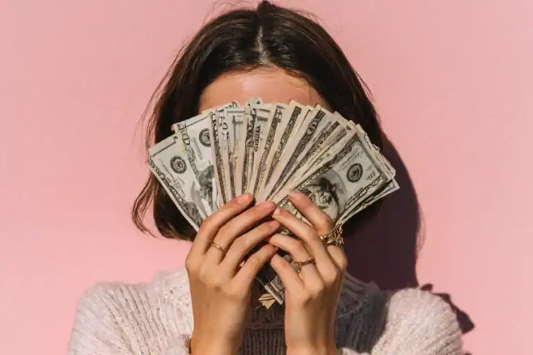 Stacks of US dollar bills held by a woman with a pink background.