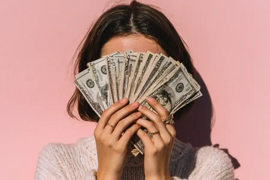 Stacks of US dollar bills held by a woman with a pink background.
