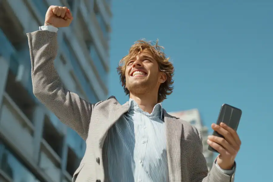 1. Happy young man celebrating success outdoors with a smartphone in hand.