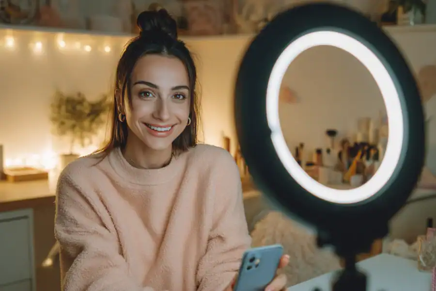 Bright young woman recording content with ring light for social media, cozy home background.