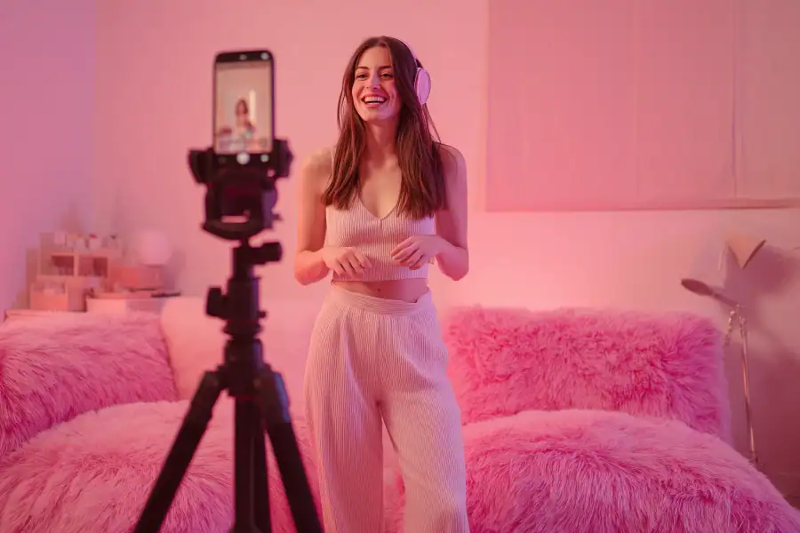 Smiling woman recording a video with a smartphone on tripod in pink-lit bedroom.